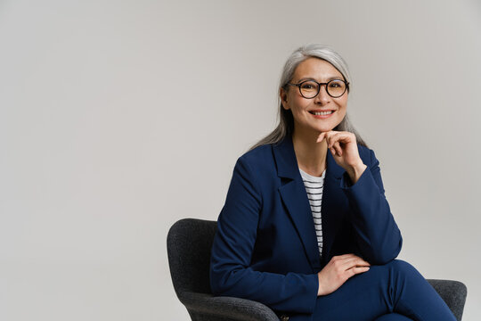 Asian mature woman wearing eyeglasses smiling while sitting in armchair