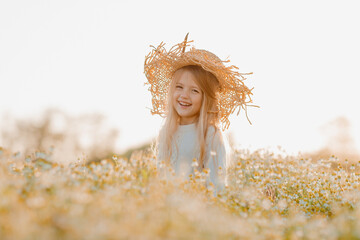 Happy little girl in a cotton dress and straw hat runs in a chamomo field in summer at sunset