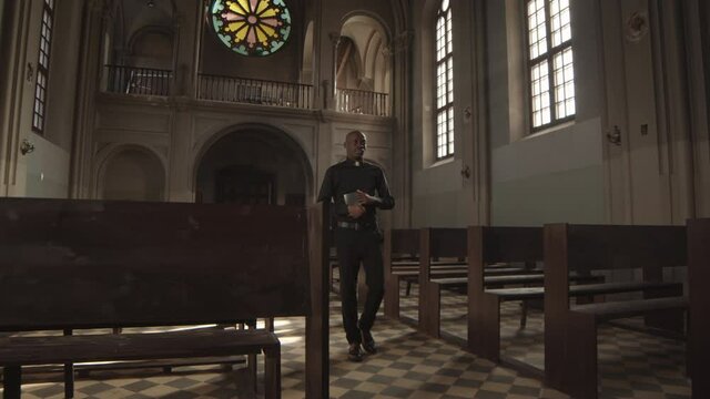 Wide shot of young African-American priest wearing black clothes and white collar going along Lutheran church with Bible in hands