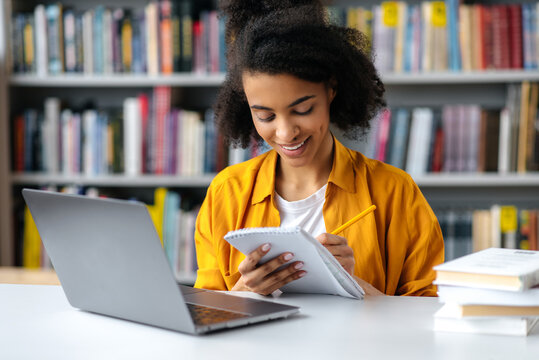 Focused satisfied successful african american female student sitting at table in a library, taking notes in notebook during lecture, gains knowledge, smiling, education concept