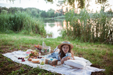 A girl in a long summer dress with long hair is on a white blanket with fruits and pastries, white basket with flowers. Concept of having picnic in a city park during summer holidays or weekends. 