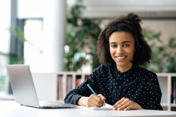 Charming successful African American young woman, in a headset, freelancer, employee or manager, sits at a work table with a laptop and a notebook, takes notes, looks at the camera, smiles friendly