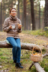 picking season, leisure and people concept - young asian woman with mushrooms in basket drinking tea and eating sandwich in autumn forest