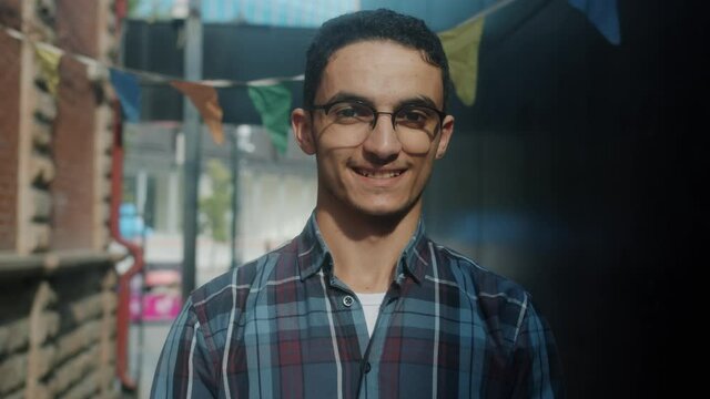 Portrait of cheerful Arab man looking at camera with happy smile standing outdoors alone wearing casual clothing and glasses. People and city concept.