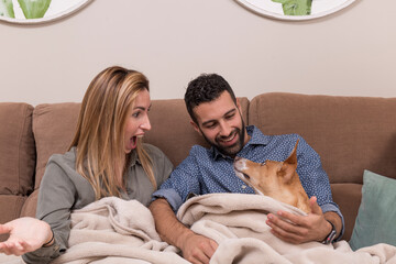 couple together with their pet, partner and their dog sitting on a sofa and covered with a blanket, the woman has an astonished face, the man smiles and the dog looks carefully at both