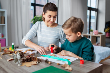 family, creativity and craft concept - mother and little son with glue sticks and paper making pictures of dry autumn leaves, pine cones and chestnuts at home