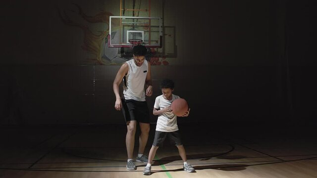 Handheld tracking shot of young male basketball player in sportswear teaching his little son how to dribble ball on dark indoor court