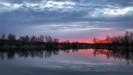 Dramatic pink sunset over forest lake with bare tree silhouettes on horizon reflected on water surface