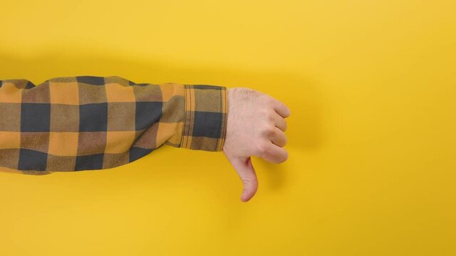 A man's hand appears on the left side of the screen and gives a thumbs down. Yellow background. The concept of disapproval and dislike.