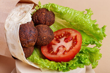 Close up of falafel pita roll with ripe red tomato and green salad in craft paper bag on beige background. Healthy vegetarian fastfood with chickpea and vegetables