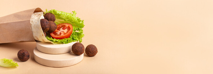 Horizontal banner with tasty falafel pita roll with ripe red tomato and green salad in craft paper bag for delivery on light circle podiun on beige background. Copy space. Healthy vegan fastfood