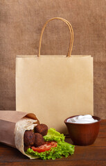 Fastfood delivery of falafel pita rollin craft paper bag. Yogurt sauce in clay bowl on brown board with burlap background. Falafel sandwich with tomato and green salad. Jewish cuisine. Place for logo