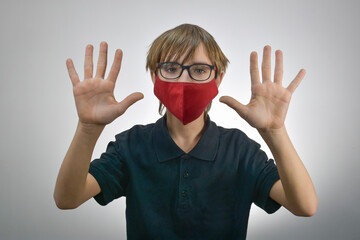 A teenager wearing glasses and a red protective medical mask with outstretched fingers on a light background with a bokeh effect. The concept of limited communication opportunities during the CAVID-19