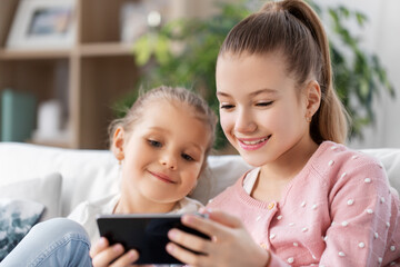 family, childhood and technology concept - two happy smiling little girls or sisters with smartphone at home