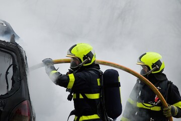 Firefighters with breathing apparatus extinguish a car that is completely engulfed in fire
