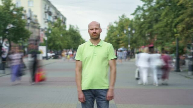 Zoom-out time lapse of pensive man standing in street downtown and looking at camera while men and women are walking around in summer. Youth and society concept.