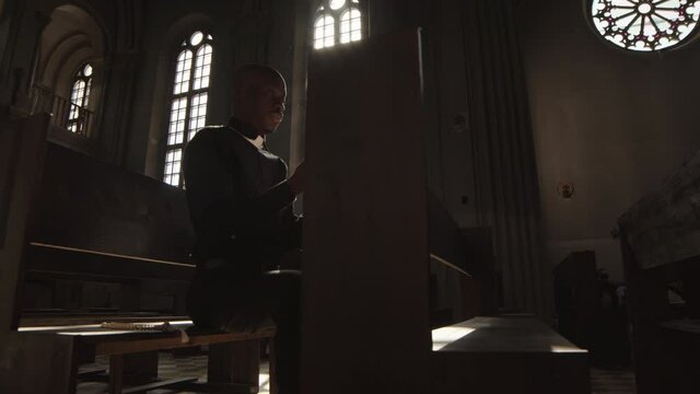 Tracking left of African-American pastor wearing black shirt and white collar sitting on wooden bench in Catholic church with Bible in hands and praying