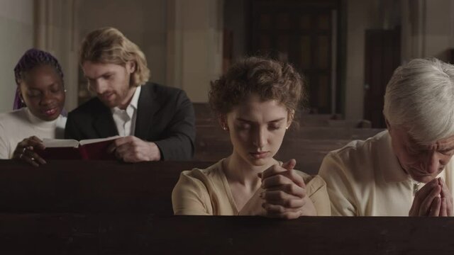 Tracking left of four parishioners of different age and race sitting on wooden benches in Lutheran church and praying. Caucasian man and African woman reading Bible on background