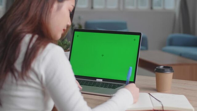 Girl Uses Laptop With Green Mock-Up Screen While Writing On Notebook In Her Living Room