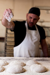 young baker in bakery shop sprinkling flour with hands on fresh bread dough in front of oven. concept of traditional manual bread preparation in bread factory