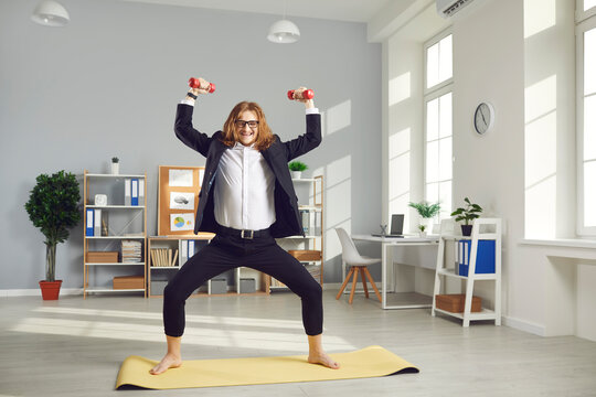 Funny nerdy barefoot employee in glasses having fitness workout during work day. Happy fit male office worker in business suit standing on gym mat and doing sports exercise with light weight dumbbells