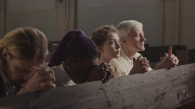 Tracking left of four parishioners of different age and race sitting on wooden bench in Lutheran church with closed eyes and hands folded for prayer and praying