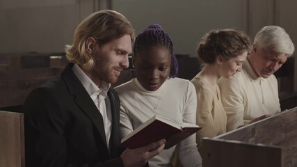 Medium shot of four parishioners of different age and race sitting on wooden bench in Lutheran church and reading Holy Bible