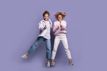 Fototapeta premium Blonde sister are jumping on a purple wall smiling and gesturing the gun sign