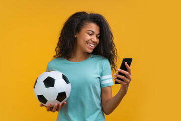 Beautiful smiling dark skinned girl holding soccer ball and watching online broadcast on his mobile phone cheering for favourite team, making bets online and waiting for winning results