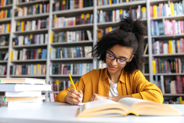Teenage african american female student studying while sits at the table in the college library, reads books to searching information for a lesson or exam, doing homework and notes, gaining knowledge - obrazy, fototapety, plakaty