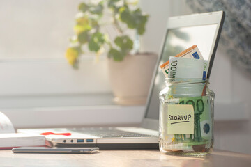 A glass jar with STARTUP written on it, with coins and banknotes inside stands on a desk.The concept is funding and promoting an innovative idea. Laptop, smartphone, diary on the desk.