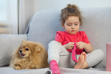A little pretty curly-haired girl sits on the couch and puts on a sock, next to her lies a dog, a German Spitz. The child learns to dress. Children development.
