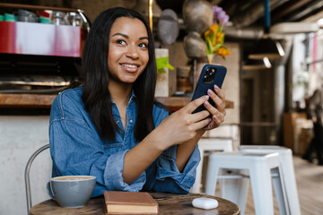 Black smiling woman using cellphone while sitting in cafe
