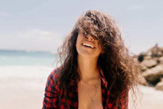 Close up outdoor portrait of adorable lovely woman with wavy hair covering face with hair smiling on the beach in sunlight
