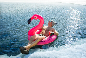 A man swimming in an ice hole in winter in Finland, floating on a pink inflatable flamingo with cocktail in hand. Vacation options, dreaming of summer. 