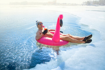 A man swimming in an ice hole in winter in Finland, floating on a pink inflatable flamingo with cocktail in hand. Vacation options, dreaming of summer. 