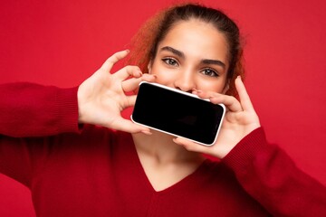 Closeup photo of young woman wearing dark red sweater isolated over red background holding smartphone and showing mobile phone screen with copy space for cutout looking at camera