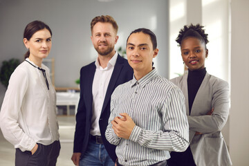Confident young man standing in office with group of coworkers. Portrait of successful male business leader, company executive manager or startup CEO with team of multiethnic employees and colleagues - obrazy, fototapety, plakaty