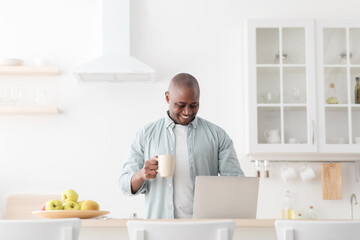Excited mature black man using laptop in kitchen and drinking coffee, browsing social media or reading online news