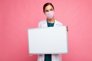 Woman doctor wearing a white medical coat and a mask holding blank board with copy space for text isolated on background. Coronavirus concept