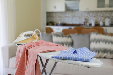 Clean freshly laundered clothes and electric iron on pressboard in living room. Pile of t-shirts and sweaters on ironing board, home interior background. Copy space, close up, top view.