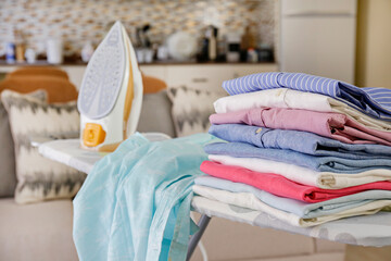 Neat stack of clean freshly laundered clothes and electric iron on pressboard in living room. Pile...