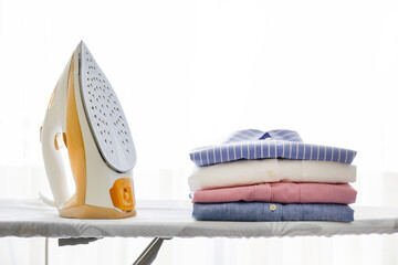 Stack of clean freshly laundered clothes and electric iron on pressboard at home. Pile of shirts on...