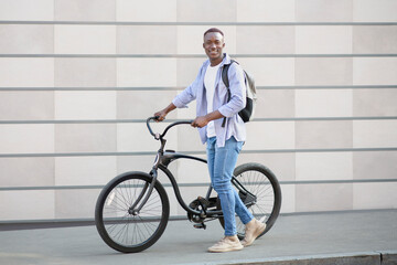 Handsome black guy with backpack and modern bike near brick wall on city street