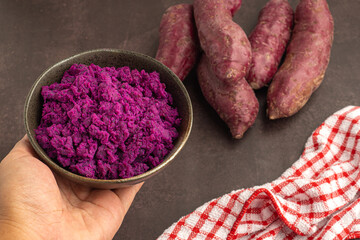 Hand holding of mashed purple sweet potatoes in a bowl with purple yams pile and a cloth placed on...