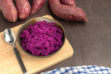 Purple yams pile and mashed purple sweet potatoes on a bowl placed on a cutting wooden board on a dark brown background. Space for text. Vintage background.  Healthy fruits concept