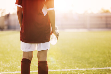 Child in Sports Uniform Holding Water Bottle in Hand. Sun Shining in Summer Day in the Background. Football Soccer Stadium and Grass Field Blurred in the Background