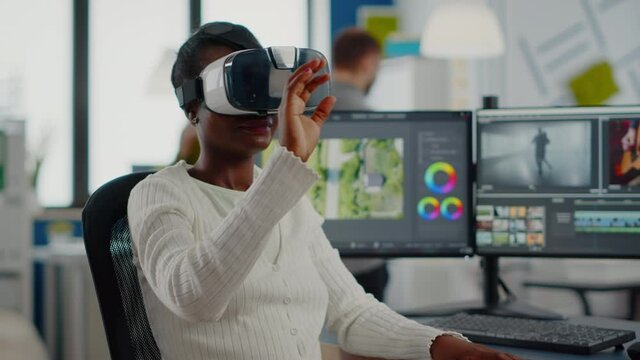 Black videographer wearing VR headset editing project in production studio office. Video maker employee processing film montage in digital multimedia company while, colleagues working in background