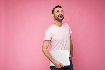 Handsome happy young unshaven man holding laptop computer looking up in t-shirt on isolated pink...