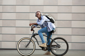 Joyful black guy with backpack and headphones riding bicycle near brick wall on city street